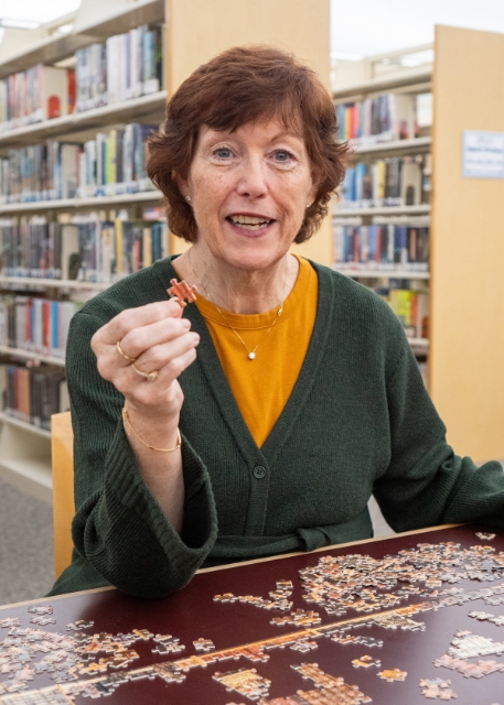 The Library Director sits at a table in a library, holding a puzzle piece and smiling, with an unfinished jigsaw puzzle in front of her.