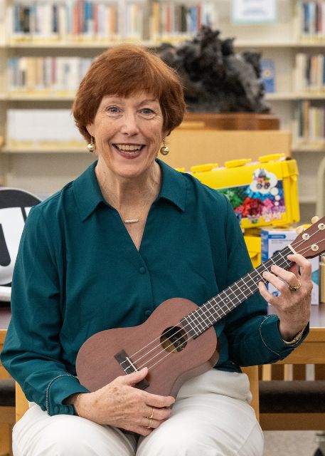 The Library Director sits at a table in a library, holding a puzzle piece and smiling, with an unfinished jigsaw puzzle in front of her.