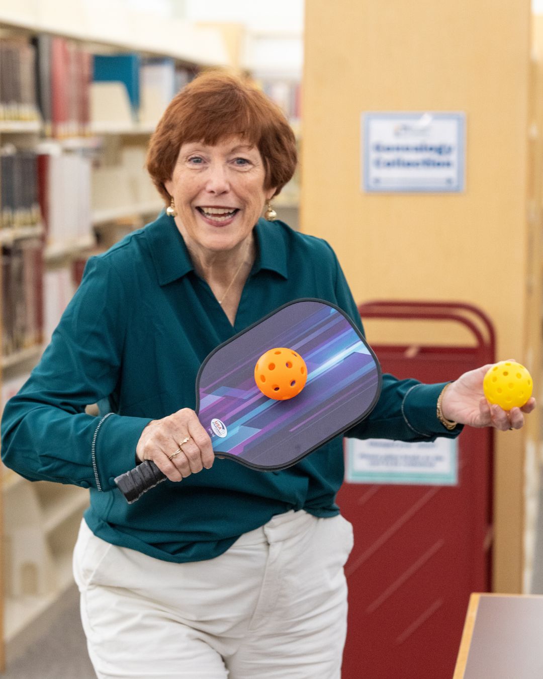 The Library Director sits at a table in a library, holding a puzzle piece and smiling, with an unfinished jigsaw puzzle in front of her.