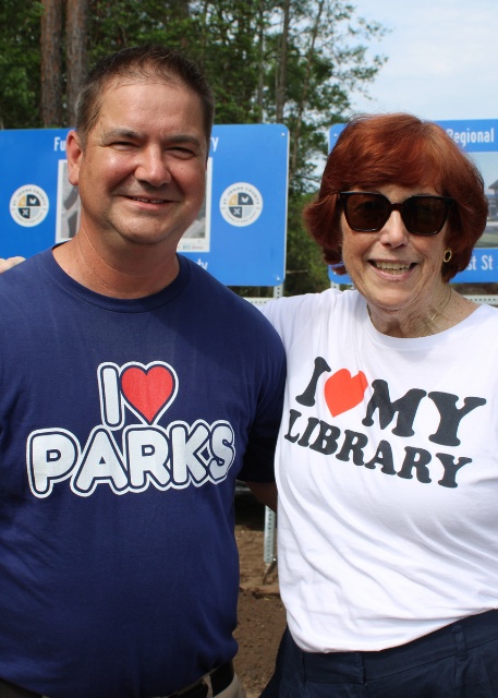 A smiling man and woman stand together outdoors, wearing shirts with heart logos reading 