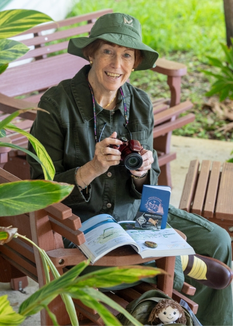 Elderly woman in a green hat and jacket sits on a bench with a camera, surrounded by plants. She smiles warmly, holding a photo book and plush toy.