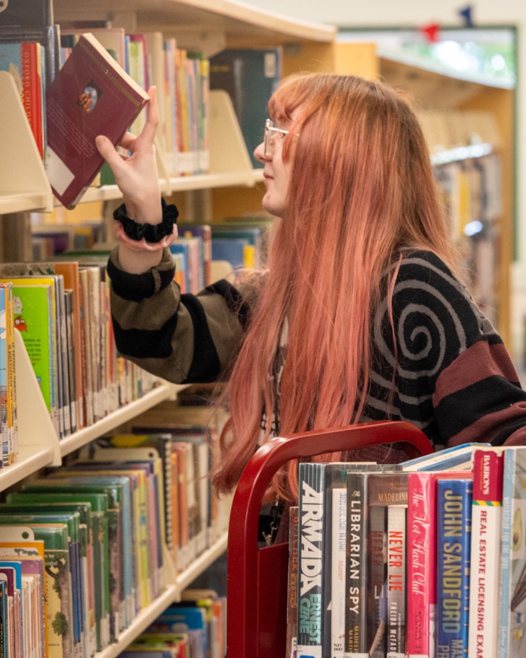 Young woman with long hair and glasses, in a library choosing a book from a shelf. She wears a striped sweater. A cart with books is nearby.
