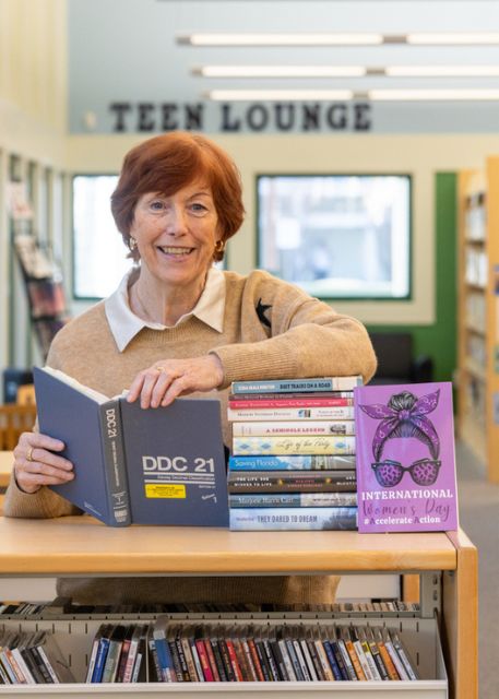The Library Director stands in a library holding a Dewey Decimal book. A stack of novels for Women's History Month are on the shelf.