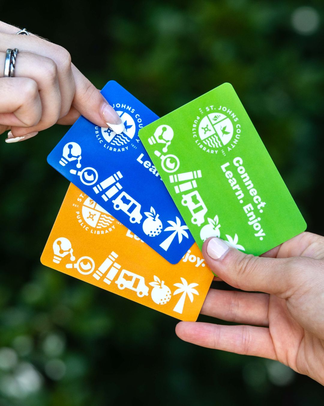 Three hands hold colorful library cards—blue, green, and orange. Each card features white icons of books, a magnifying glass, and a palm tree, conveying learning and community.