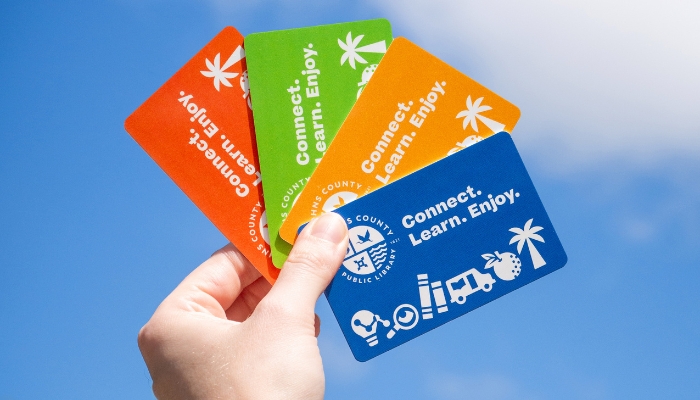 A hand holds four colorful library cards against a blue sky. The cards are yellow, green, orange, and blue, each with the text 