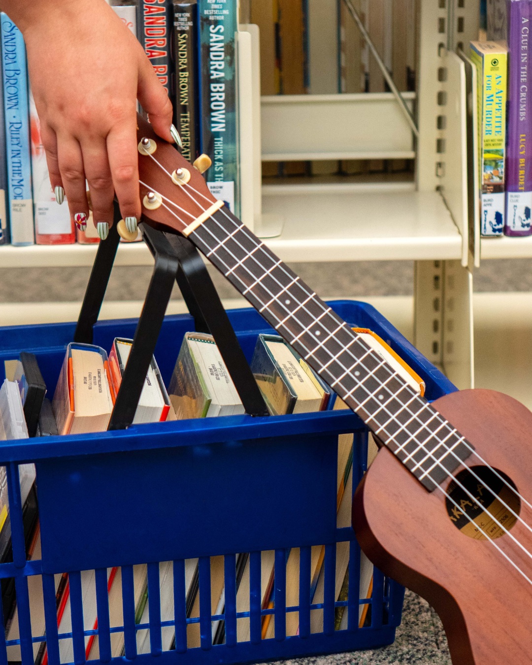 A hand lifts a blue basket filled with books and a ukulele in front of a library bookshelf.