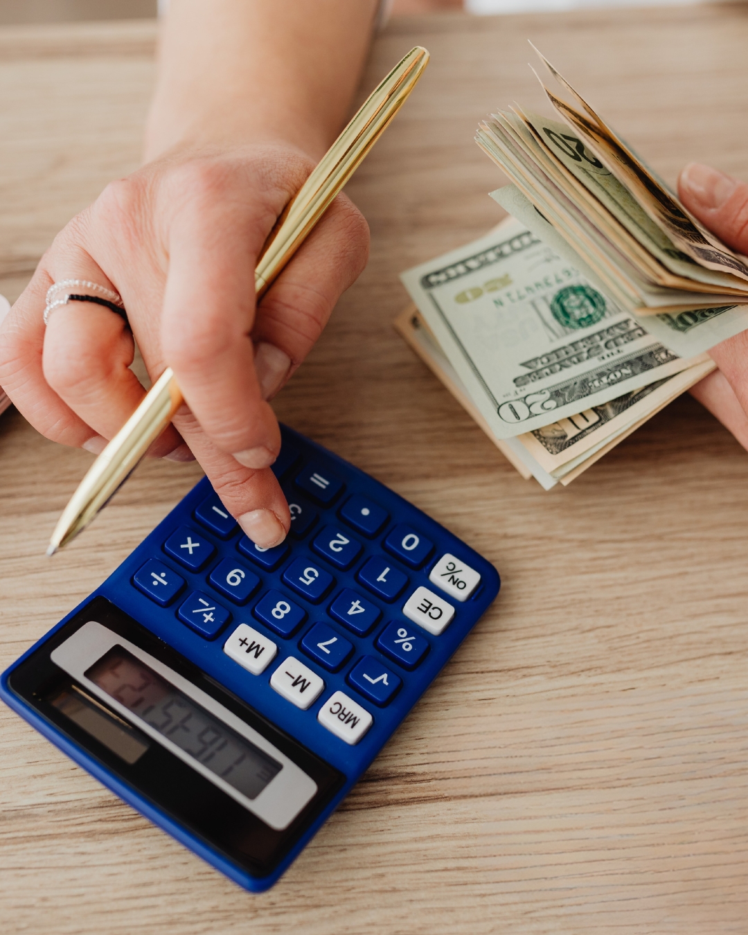 Hands using a blue calculator and counting a stack of US dollar bills on a wooden table, conveying a sense of budgeting and financial planning.