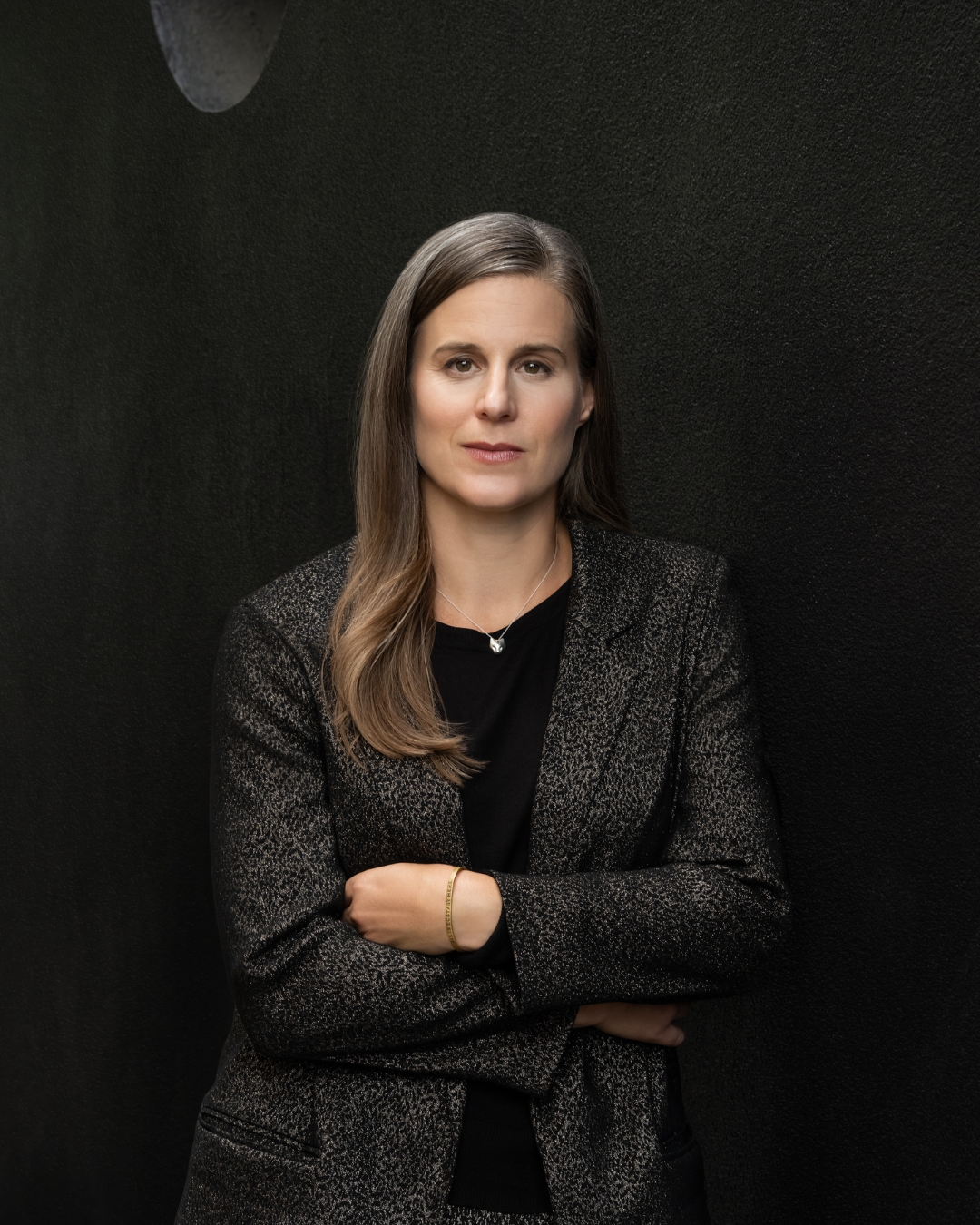 Professional portrait of a confident woman with long brown hair in a textured blazer, arms crossed against a black backdrop.