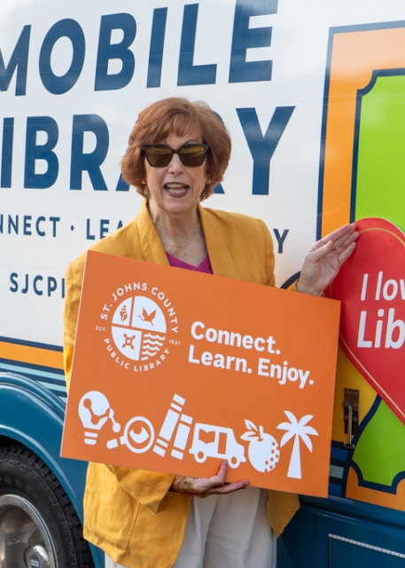 Woman in sunglasses smiles beside a mobile library van, holding an orange St. Johns County library sign reading “Connect. Learn. Enjoy.”