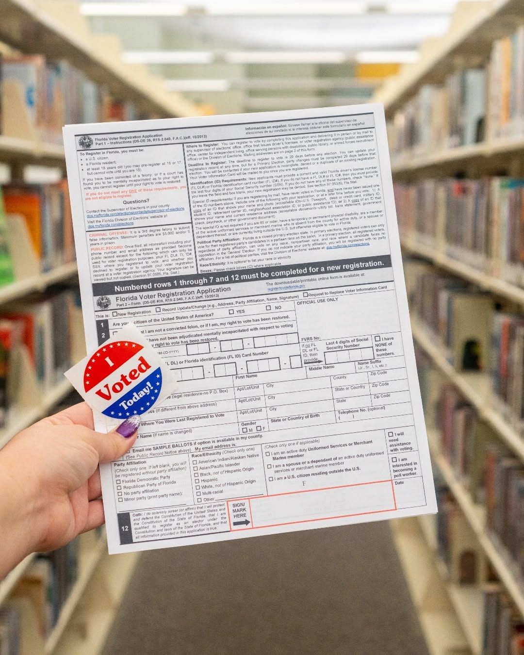 Hand holds a voter registration form with a red, white, and blue “I Voted Today!” sticker in a library aisle, conveying civic participation.