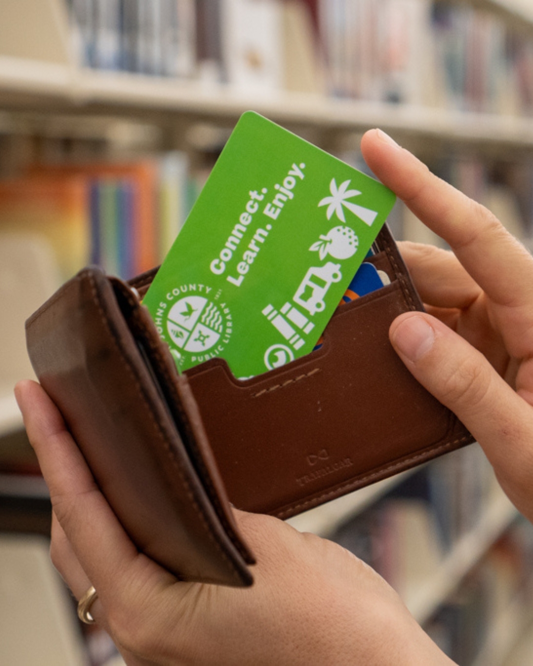 Hands hold an open brown leather wallet with a bright green “Connect. Learn. Enjoy.” card, against a softly blurred bookstore shelf.