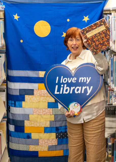 The Library Director stands in a library aisle, smiling and holding a book and a heart-shaped sign reading 